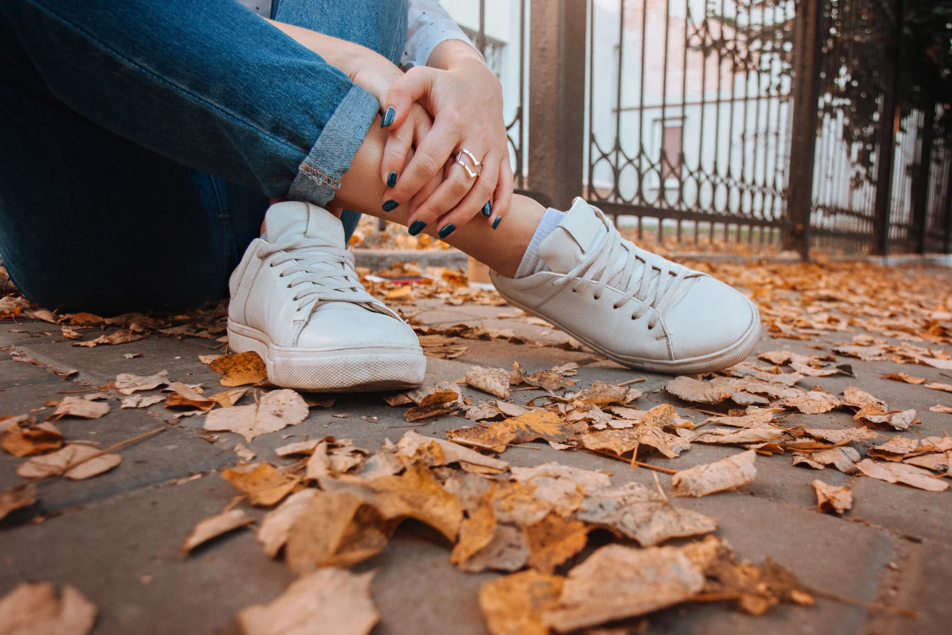 Zbliżenie na kobietę siedzącą na chodniku w białych sneakersach, otoczoną opadłymi jesiennymi liśćmi.